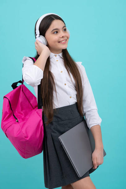 Back to school. Teenager schoolgirl in school uniform with bagpack, headphones and laptop. School children on isolated blue background - Фото, зображення