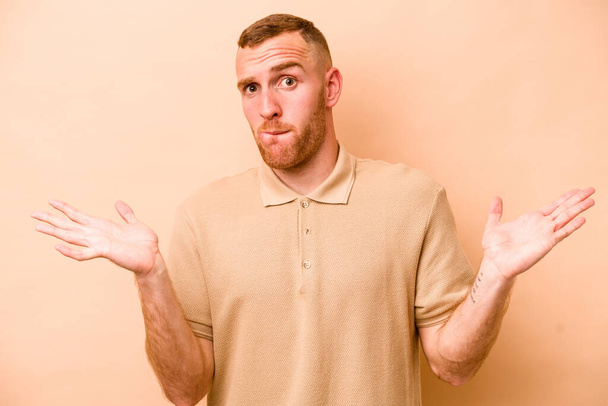 Young caucasian man isolated on beige background confused and doubtful shrugging shoulders to hold a copy space. - Photo, Image
