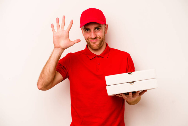 młody dostawy kaukaski mężczyzna gospodarstwa pizze odizolowany na biały tło uśmiechnięty wesoły pokazując numer pięć z palce. - Zdjęcie, obraz