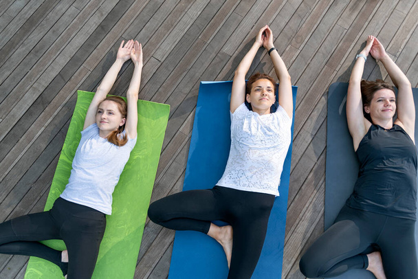 Een groep jonge vrouwen die buiten yoga doen. Drie meisjes liggen op yoga matten en voeren asana - Foto, afbeelding