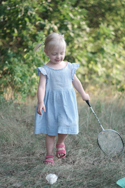 Little girl in blue dress playing badminton in the park. Blonde girl with racket outside. Vertical frame - Foto, Imagen