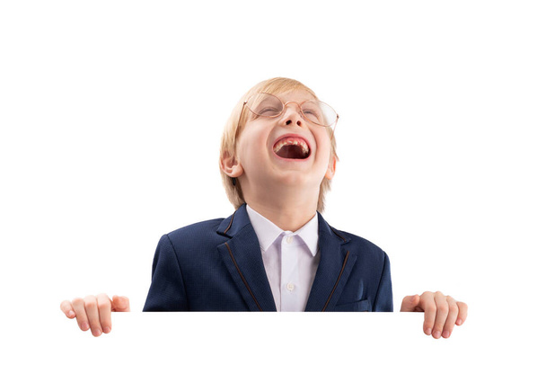 Χαρούμενο αγόρι ποζάρει με άδειο λευκό χάρτινο πίνακα και γελάει. Χαριτωμένο ξανθό αγόρι κρυφοκοιτάζει από πίσω από το λευκό πίνακα - Φωτογραφία, εικόνα