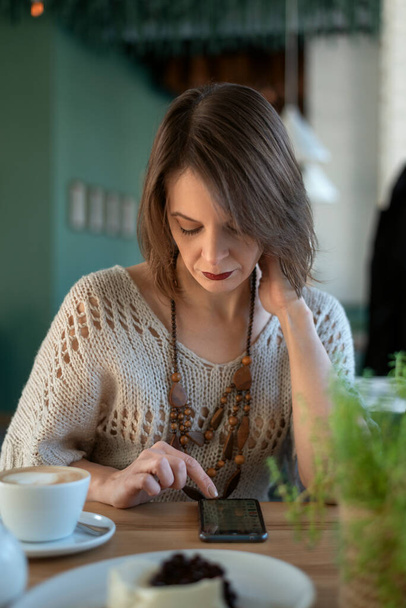 Ritratto di donna matura in caffè con telefono. Donna di mezza età si siede in un caffè con latte o cappuccino caffè e utilizza smartphone - Foto, immagini