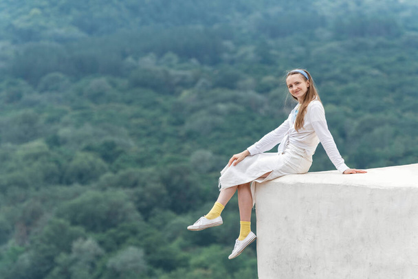 Sorrindo jovem mulher senta-se na varanda com as pernas penduradas para baixo e aponta para a distância no fundo das montanhas - Foto, Imagem