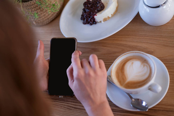 Las manos de mujer con teléfono inteligente en la cafetería contra la mesa capuchino café y postre. Smartphone en manos femeninas. Trabajo durante el almuerzo. - Foto, imagen
