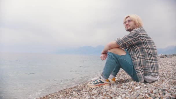Smiling young man in a plaid shirt sits on the shore. Mid shot - Footage, Video