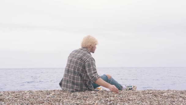Młody blondyn w koszuli w kratę siedzi na brzegu i wrzuca kamienie do morza. Środek strzału - Materiał filmowy, wideo