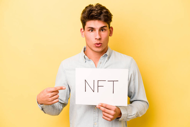 Giovane uomo caucasico in possesso di cartello NFT isolato su sfondo giallo che punta verso il lato - Foto, immagini