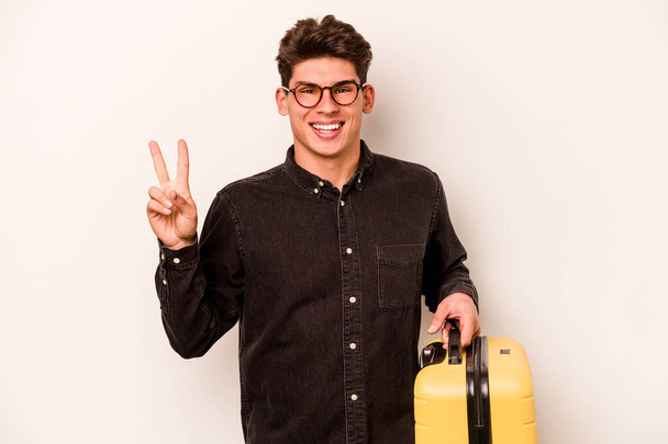 młody podróżnik biały mężczyzna trzymając walizkę odizolowany na białym tle radosny i beztroski pokazując symbol pokoju z palcami. - Zdjęcie, obraz