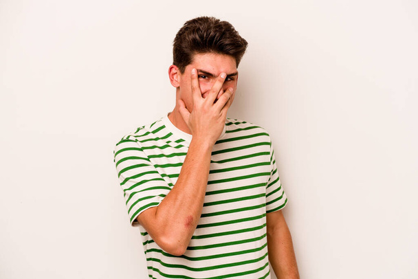 Young caucasian man isolated on white background blink at the camera through fingers, embarrassed covering face. - Photo, Image