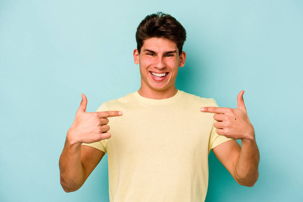 Young caucasian man isolated on blue background surprised pointing with finger, smiling broadly. - Photo, Image