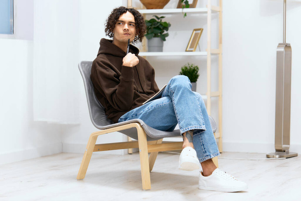 Kıvırcık saçlı bir adam not defteri ve kalem ışığı arkaplanı olan bir sandalyeye oturur. Yüksek kalite fotoğraf - Fotoğraf, Görsel