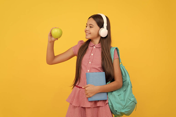 Ημέρα γνώσης. έννοια της εκπαίδευσης. Παιδί με ακουστικά με βιβλίο και μήλο για μεσημεριανό. Σεπτέμβριος 1. Υγιής παιδική ηλικία. Παιδί με σχολική τσάντα. Μουσική. Χαρούμενη έφηβη κουβαλάει σακίδιο. Πίσω στο σχολείο.. - Φωτογραφία, εικόνα