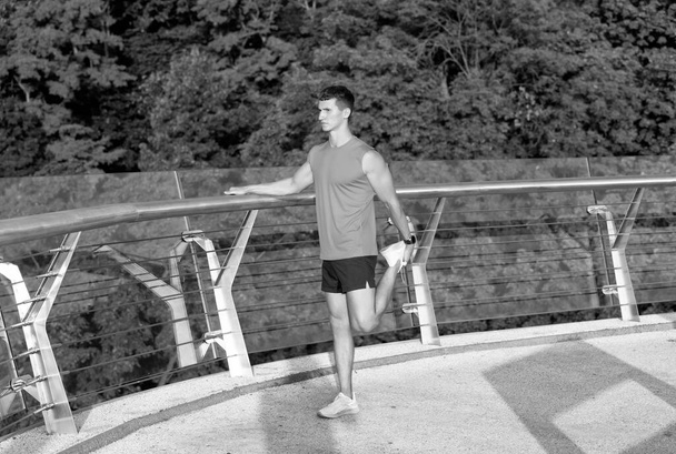 Athletic guy in sportswear grab ankle with hand stretching leg after outdoor training on promenade, stretch. - Foto, immagini