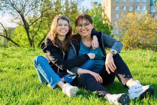 Happy mother and daughter hugging sitting on grass, looking at camera. Mom and girl resting together on lawn. Family, happiness, leisure, lifestyle, relationship love, mothers day, motherhood concept - Foto, Imagen