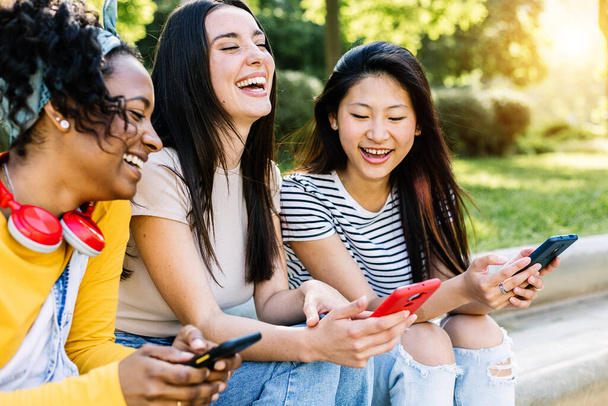 Felices amigas jóvenes multirraciales que se divierten usando teléfonos móviles en la calle: tres mujeres se ríen juntas mientras ven contenido divertido de redes sociales en la aplicación para teléfonos inteligentes - Foto, imagen