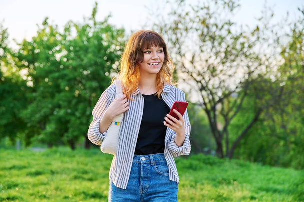 Outdoor portrait of trendy young red-haired girl with smartphone in hand, green trees sky background. Smiling attractive teenage female student. Youth, beauty, fashion, lifestyle, leisure concept - Photo, Image