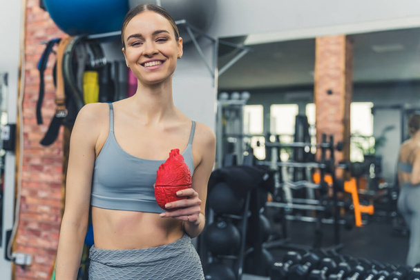 Jeune femme blanche mince excitée debout dans une salle de gym bien équipée, souriant à la caméra, tenant un faux cœur humain rouge. Photo de haute qualité - Photo, image