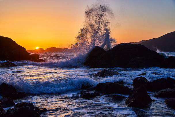 Bild von einer großen Welle, die bei herrlichem Sonnenuntergang gegen Felsen am Strand kracht - Foto, Bild