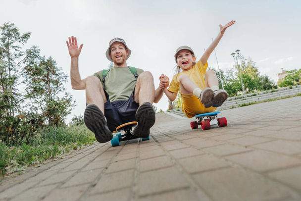 Glücklicher Vater und seine Tochter sitzen auf Skateboards im Freien. Verspielte junge Mann und Vorschulmädchen genießen Spaß zusammen im Sommer. Konzept für Familienaktivitäten. - Foto, Bild
