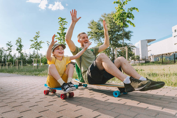 Šťastný otec a jeho dcera sedí na skateboardech venku. Hravý mladý muž a školačka dívka těší bavit čas spolu v létě. Koncept rodinných aktivit. - Fotografie, Obrázek