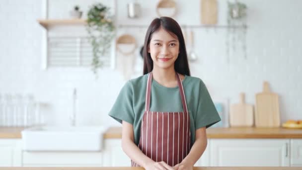 Onnellinen nuori aasialainen nainen yllään esiliina ja seisoo keittiössä huoneessa. Kaunis nainen hymyilee ja katselee kameraa luottavainen olo - Materiaali, video