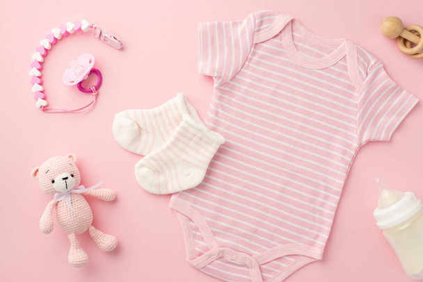 Концепция детских принадлежностей. Вид сверху фото розовой детской одежды боди носки соска цепи бутылка деревянная погремушка и вязаные игрушки плюшевый медведь на изолированном розовом фоне пастель - Фото, изображение