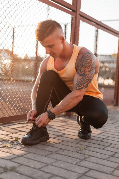 Handsome young stylish man with hairstyle and maya tattoo in trendy t-shirt, shorts and leggings ties laces on black sneakers at sunset on the street - Fotoğraf, Görsel