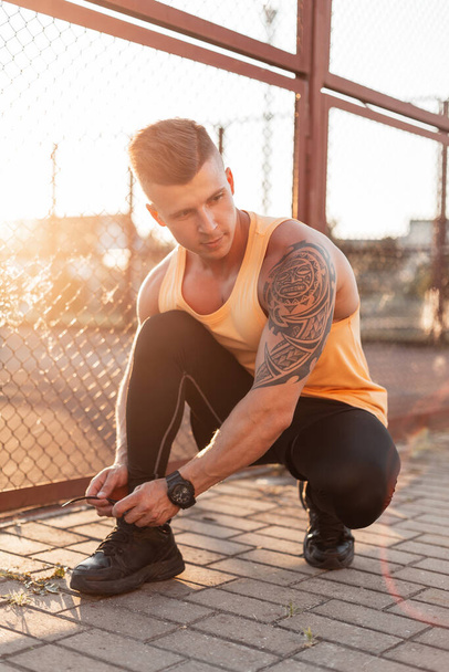 Μοντέρνος όμορφος νεαρός άνδρας με χτένισμα και τατουάζ στον ώμο του δένει τα κορδόνια του σε αθλητικά παπούτσια στο δρόμο το ηλιοβασίλεμα - Φωτογραφία, εικόνα