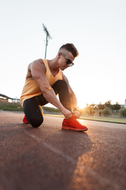 Молодой красивый спортсмен в красных кроссовках завязывает шнурки и готовится бежать на закате. Спорт и здоровый образ жизни - Фото, изображение