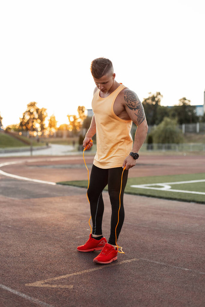 Handsome fitness young male athlete in fashionable sports clothes with red sneakers with a skipping rope at the stadium. Athletic guy getting ready for exercise and workout at sunset - Photo, Image