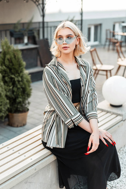 Красивая молодая девушка-подросток с светлыми волосами в модной одежде с рубашкой, топом, юбкой и очками сидит на скамейке в городе - Фото, изображение