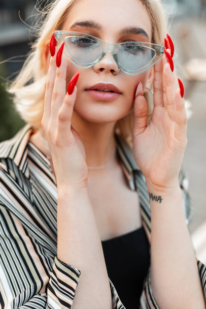 Fashion portrait of young tinder blonde woman puts on stylish blue sunglasses in the city - Photo, Image