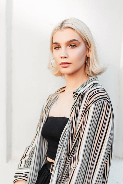 Fashion young girl with blonde short hair in stylish summer shirt and black top stands near a white wall - 写真・画像