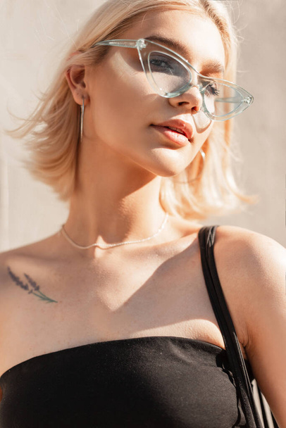 Vogue portrait de belle jeune adolescente blonde modèle féminin avec apparence adorable avec des lunettes bleues à la mode près de fond blanc à l'extérieur par une journée ensoleillée. Jolie femme - Photo, image