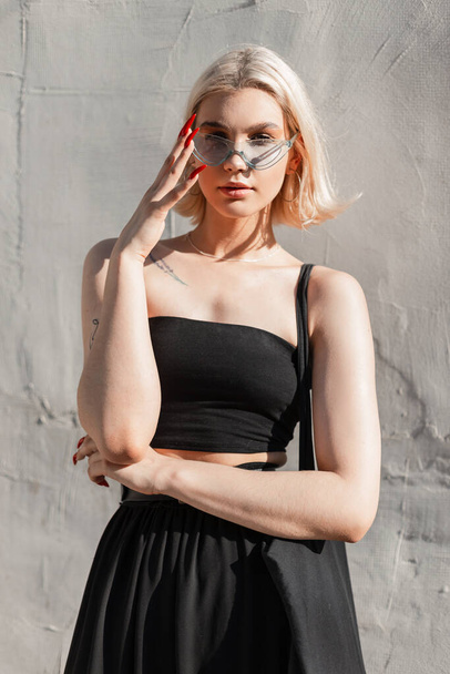 Cool pretty young stylish girl with trendy blue glasses in a fashion black top and skirt with bag stands near a street wall and looks at the camera - Fotografie, Obrázek
