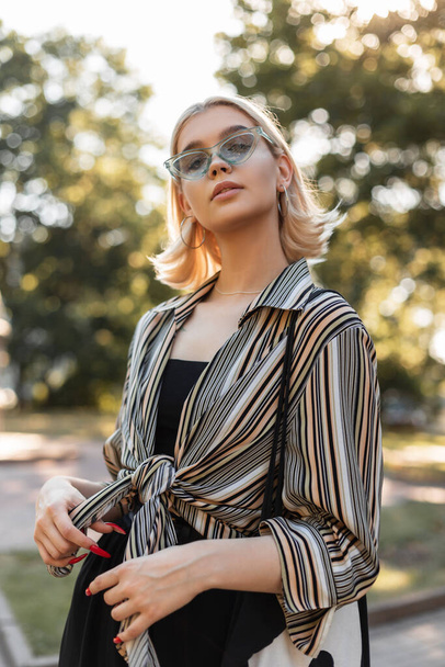 Pretty cool young woman with trendy sunglasses in vintage shirt and black dress with bag walks outdoors in sunny summer day. Female spring fashion look - Φωτογραφία, εικόνα