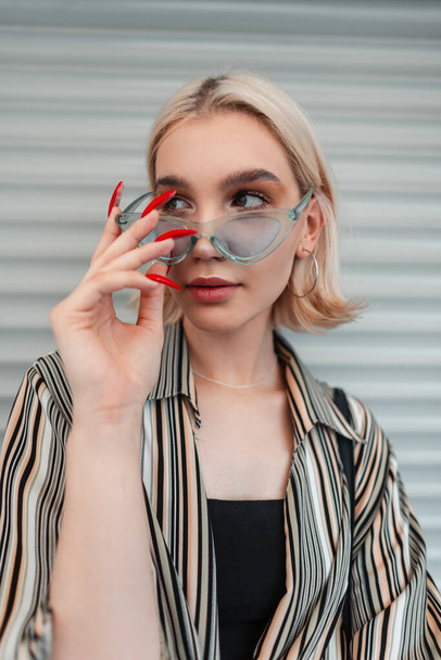Cool fashion young woman with long nails in trendy shirts puts on blue summer glasses and walks on the street near a metal wall - Фото, изображение