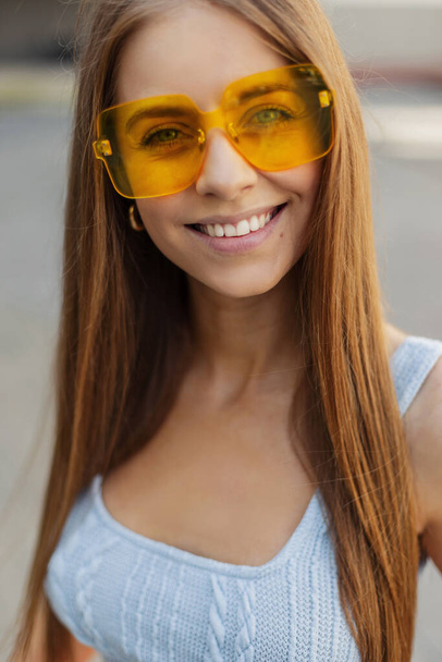 Beautiful young happy model girl with a cute smile in a fashionable knitted tank top with colorful fashion sunglasses looks at the camera in the city - Photo, image