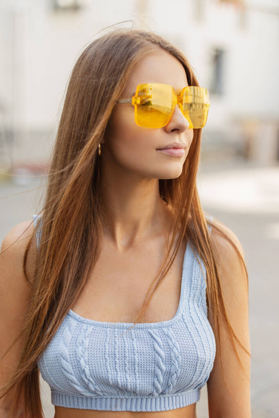 Hermosa mujer elegante con gafas de sol de color amarillo brillante con un elegante top de punto azul de verano y caminar por la ciudad. Estilo de ropa de color femenino look - Foto, Imagen