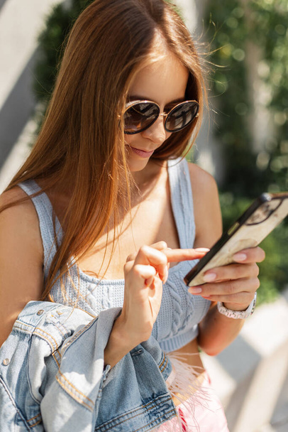 Belle jeune fille hipster élégant avec des lunettes de soleil cool écrit un message et utilise un smartphone dans la rue - Photo, image