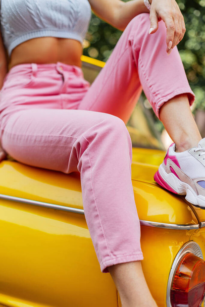 Pembe kotlu, şık bacakları ve sarı renkli spor ayakkabıları olan şık bir kadın sarı bir arabanın üzerinde oturuyor. Kadınların yaz tatili, parlak moda. - Fotoğraf, Görsel