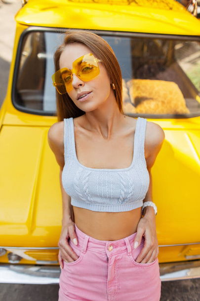 Sexy belle femme lumineuse avec des lunettes de soleil jaunes élégantes dans un haut d'été à la mode et un jean rose à la mode se trouve à côté d'une voiture jaune vintage rétro par une journée ensoleillée, vue sur le dessus. Vacances d'été et positivité - Photo, image