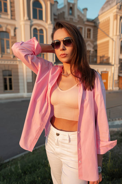 Cool trendy beautiful girl with fashion sunglasses in pink shirt with tank top walks in the city. Urban glamour female style look - Photo, Image