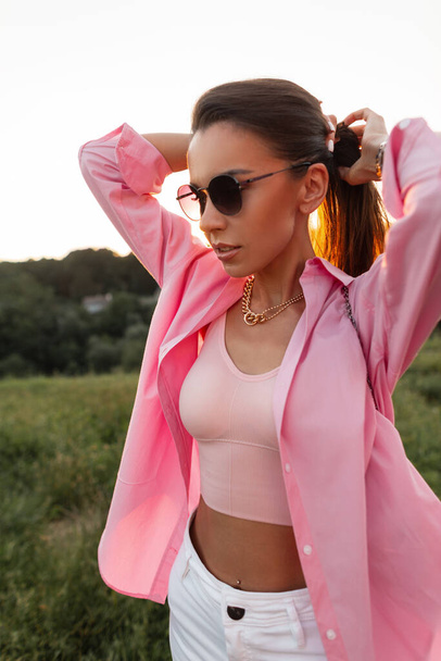 Glamorous female portrait of beautiful girl with trendy round sunglasses in pink fashionable shirt and tank top walks outdoors - Foto, afbeelding