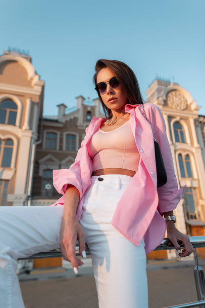 Stylish beautiful young girl with glasses in a fashionable pink shirt and tank top with handbag sitting in the city at sunset on a summer day - 写真・画像