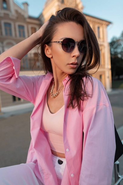 Élégant beau modèle jeune fille dans une chemise rose à la mode et haut avec des lunettes de soleil marchant dans la ville par une chaude journée de printemps - Photo, image
