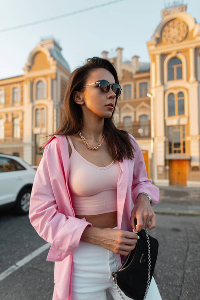Fashionable model hipster beauty girl with sunglasses in a trendy pink shirt and top zippers up her fashion purse and walking in the city - Фото, изображение