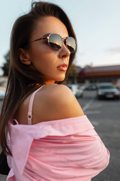 Summer female portrait of a beautiful young girl with cool stylish sunglasses in fashionable pink outfit with a shirt and bra top walking in the city at sunset - Photo, Image