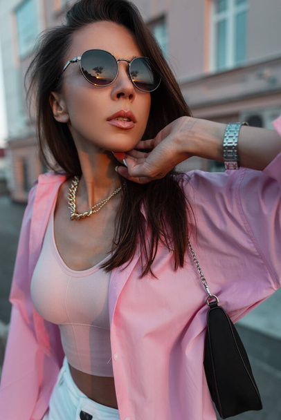 Portrait féminin élégant d'une belle jeune fille glamour avec des lunettes de soleil vintage rondes dans une chemise rose à la mode et haut avec un sac à main et une chaîne en or marchant et posant dans la ville au coucher du soleil - Photo, image
